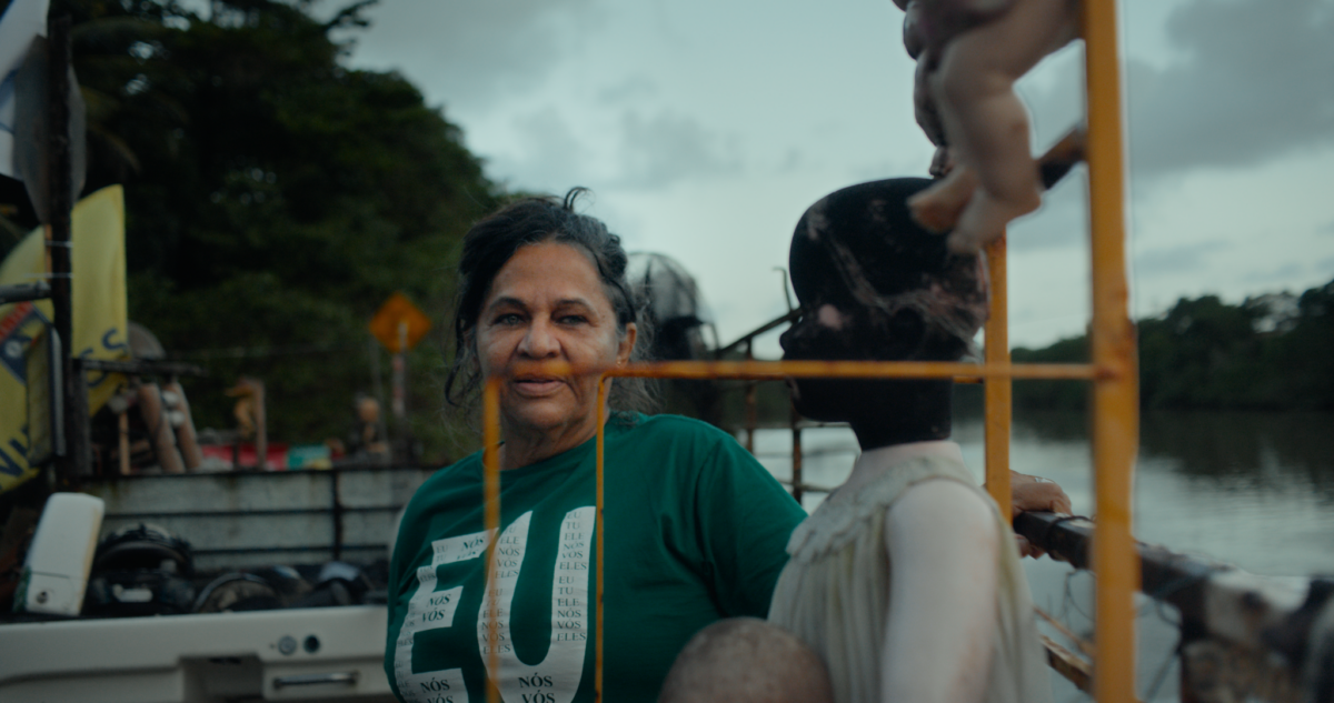 Frame do documentário Guardiãs do Futuro. Em uma paisagem às margens de um rio, uma mulher de cabelos grisalhos presos para trás veste uma camiseta verde com letras brancas. Ela está em uma embarcação ou estrutura flutuante próxima à água. À frente dela aparece uma escultura ou boneco humanoide de cor escura. Ao fundo vê-se o rio, vegetação densa e céu nublado. Elementos amarelos da estrutura criam linhas verticais e horizontais na composição. A paleta mistura verde, azul, cinza e amarelo, reforçando a relação entre comunidade e ambiente aquático.