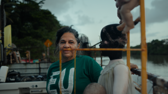 Frame do documentário Guardiãs do Futuro. Em uma paisagem às margens de um rio, uma mulher de cabelos grisalhos presos para trás veste uma camiseta verde com letras brancas. Ela está em uma embarcação ou estrutura flutuante próxima à água. À frente dela aparece uma escultura ou boneco humanoide de cor escura. Ao fundo vê-se o rio, vegetação densa e céu nublado. Elementos amarelos da estrutura criam linhas verticais e horizontais na composição. A paleta mistura verde, azul, cinza e amarelo, reforçando a relação entre comunidade e ambiente aquático.