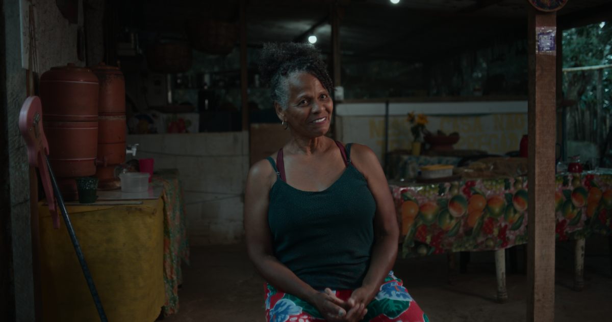 Frame do documentário Guardiãs do Futuro. A imagem tem tons escuros e iluminação quente. No centro, uma mulher negra de cabelos grisalhos presos em coque alto está sentada e sorri para a câmera. Ela veste uma regata verde-escura e uma saia estampada em vermelho, azul e branco. A cena acontece no interior de uma casa simples de madeira. À esquerda há um filtro de barro sobre um móvel amarelo, copos e utensílios domésticos. Ao fundo aparecem uma mesa coberta por toalha estampada com frutas, recipientes e objetos de cozinha. A iluminação cria um ambiente acolhedor e intimista.