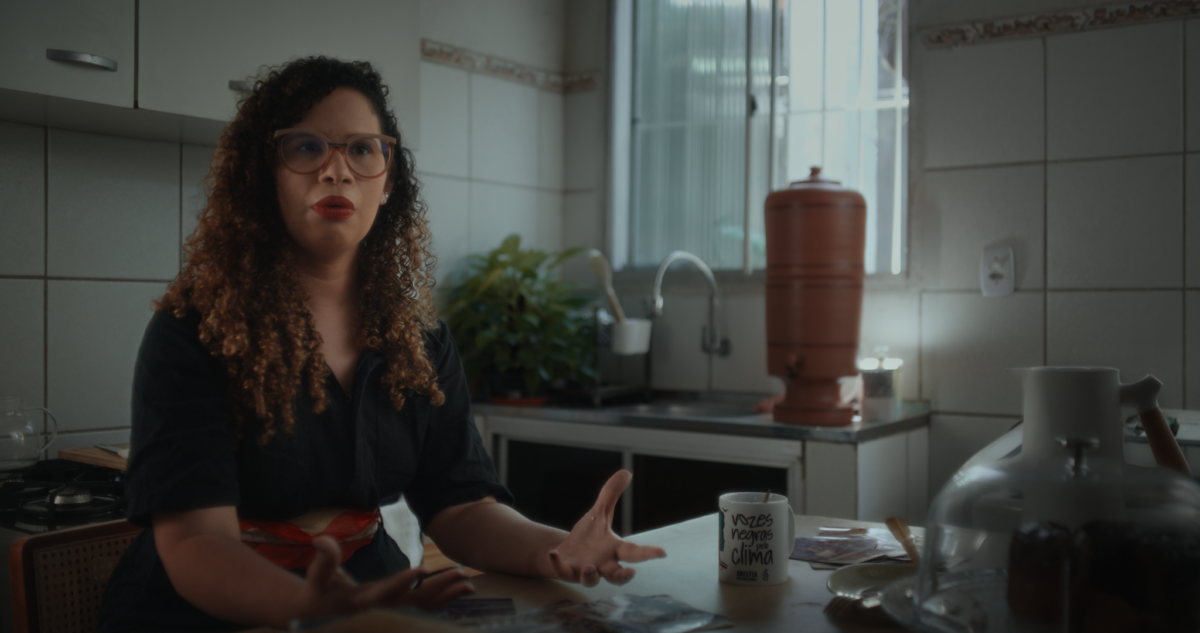 Frame do documentário Guardiãs do Futuro. Uma mulher de pele clara, cabelos cacheados longos e óculos de armação transparente está sentada à mesa de uma cozinha enquanto fala e gesticula com as mãos. Ela veste uma camisa preta. Sobre a mesa há fotografias espalhadas, uma caneca branca com a frase “Vozes negras pelo clima”, pratos e recipientes de vidro. Ao fundo aparecem uma pia, uma janela iluminada por luz natural, uma planta verde e um filtro de barro. A cena tem tons suaves de cinza, bege e verde, com iluminação que entra pela janela e destaca o ambiente doméstico.
